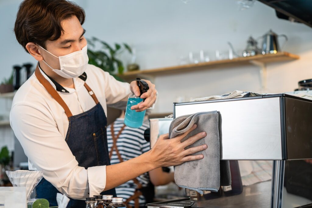 Asian attractive cafe shop owner clean counter bar and wiping on machine with sanitizer spray.
