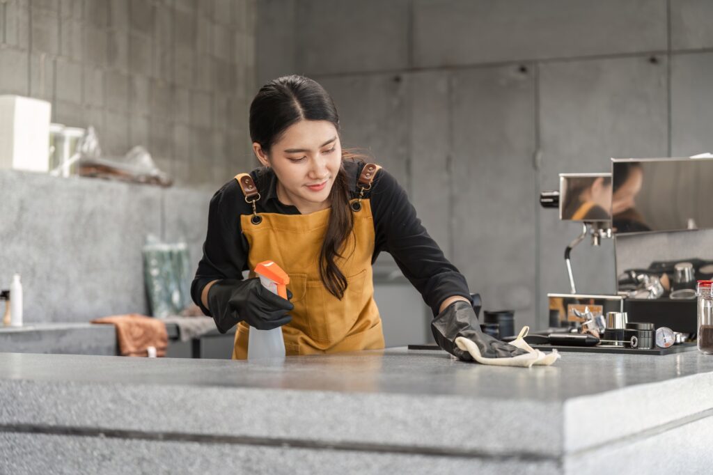 Female Employee Cleaning Modern Coffee Shop Counter with Spray Bottle and Cloth in Industrial Style